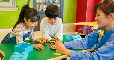 Two children and an adult sit at a green table, smiling and playing with colorful LEGO bricks, with a bookshelf and bright green walls in the background.
