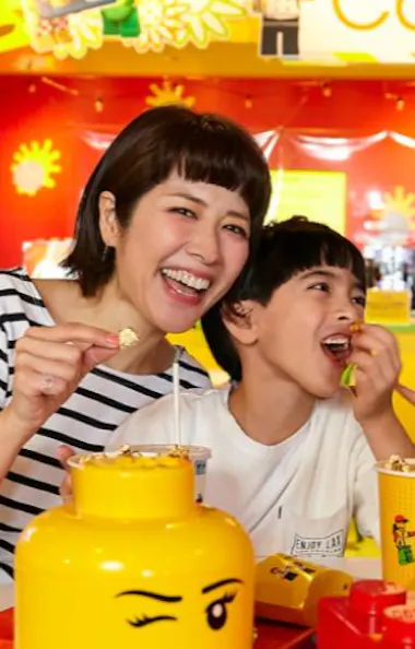 Tokyo Legoland Discovery Center A smiling family of four enjoys snacks together at a brightly colored café decorated with LEGO-themed items, including a yellow LEGO head container and red LEGO bricks on the table.