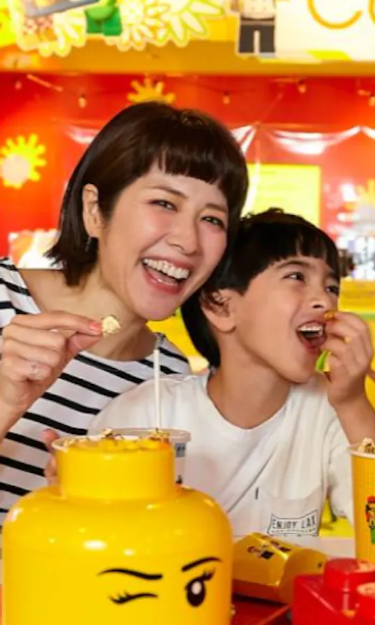 Tokyo Legoland Discovery Center A smiling family of four enjoys snacks together at a brightly colored café decorated with LEGO-themed items, including a yellow LEGO head container and red LEGO bricks on the table.