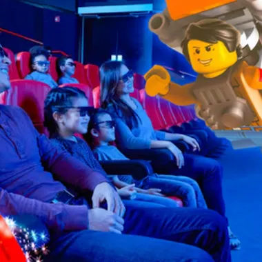 Tokyo Legoland Discovery Center A group of people sit in red theater seats, wearing 3D glasses and smiling as they watch a large animated LEGO character appear to come out of the movie screen toward them.