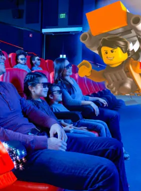 A group of people sit in red theater seats, wearing 3D glasses and smiling as they watch a large animated LEGO character appear to come out of the movie screen toward them.