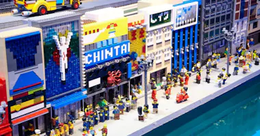 A colorful LEGO city scene with detailed buildings, storefronts, and many LEGO mini-figures walking along a busy sidewalk next to a street.