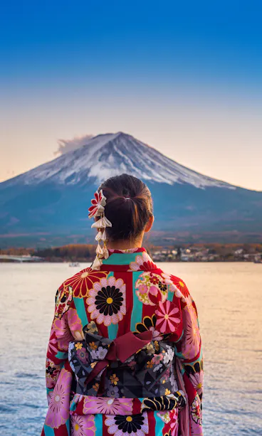 Mount Fuji View A person wearing a colorful floral kimono stands by a lake, facing Mount Fuji in the distance under a clear blue sky at sunset.