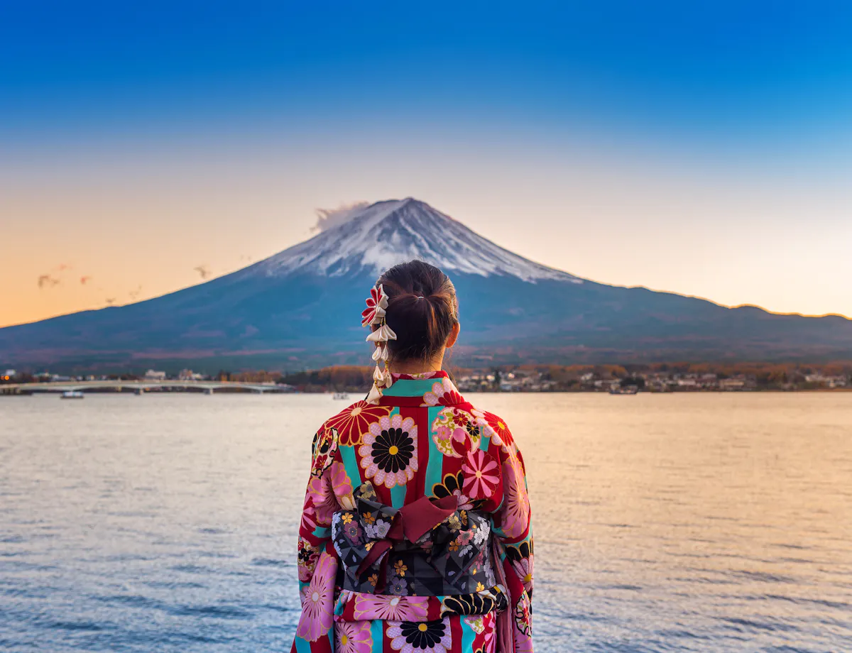 Mount Fuji View A person wearing a colorful floral kimono stands by a lake, facing Mount Fuji in the distance under a clear blue sky at sunset.