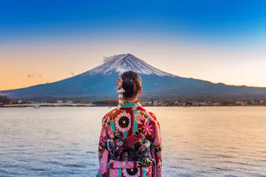 A person wearing a colorful floral kimono stands by a lake, facing Mount Fuji in the distance under a clear blue sky at sunset.