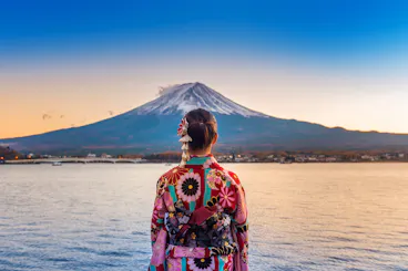 A person wearing a colorful floral kimono stands by a lake, facing Mount Fuji in the distance under a clear blue sky at sunset.