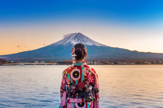 A person wearing a colorful floral kimono stands by a lake, facing Mount Fuji in the distance under a clear blue sky at sunset.