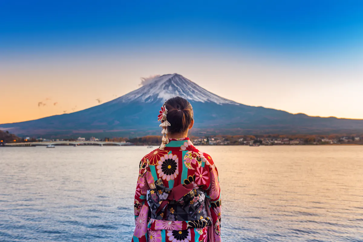 A person wearing a colorful floral kimono stands by a lake, facing Mount Fuji in the distance under a clear blue sky at sunset.