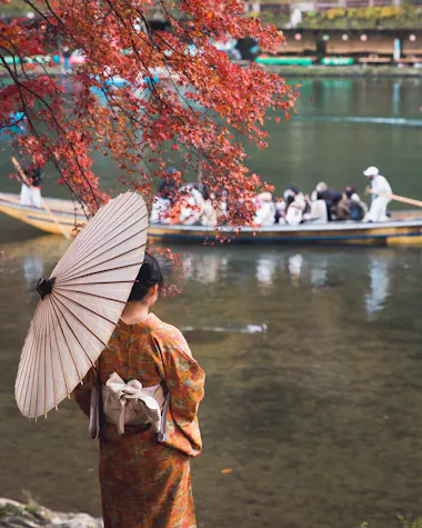 A person in a patterned kimono holds a traditional paper umbrella while standing by a river, with red autumn leaves overhead and a boat full of people on the water in the background.