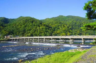 A wooden bridge stretches over a river with gentle rapids, surrounded by lush green hills under a clear blue sky.