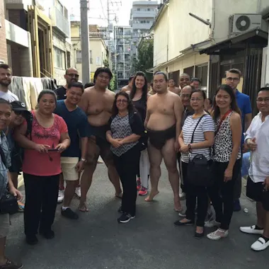 Sumo Wrestlers A group of people poses for a photo outside in an alleyway; two sumo wrestlers in traditional attire stand barefoot among casually dressed men and women, with buildings and power lines in the background.