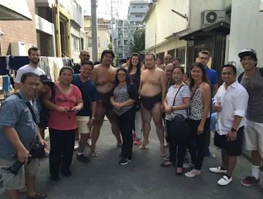A group of people poses for a photo outside in an alleyway; two sumo wrestlers in traditional attire stand barefoot among casually dressed men and women, with buildings and power lines in the background.