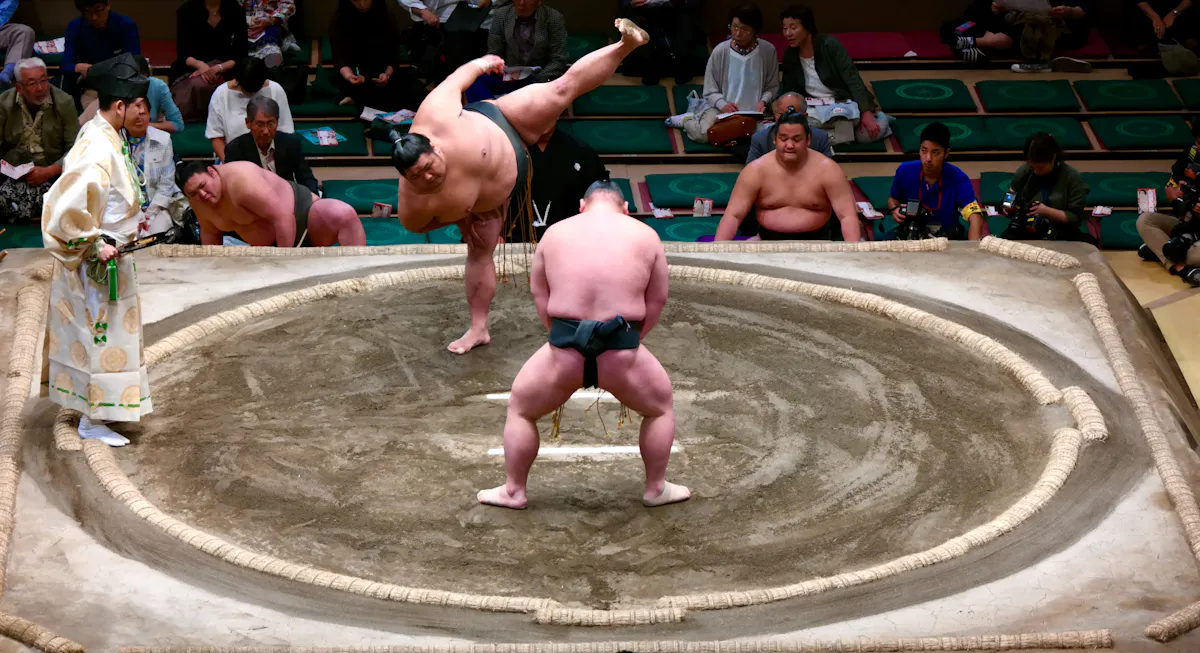 Two sumo wrestlers compete in a ring as one lifts his leg high before charging, surrounded by other wrestlers, officials, and audience members watching the match.
