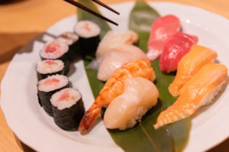 A plate of assorted sushi is arranged on a leaf. The assortment includes maki rolls, and various pieces of nigiri topped with shrimp, salmon, tuna, and other types of fish. A pair of chopsticks is poised above the sushi, ready to pick a piece.