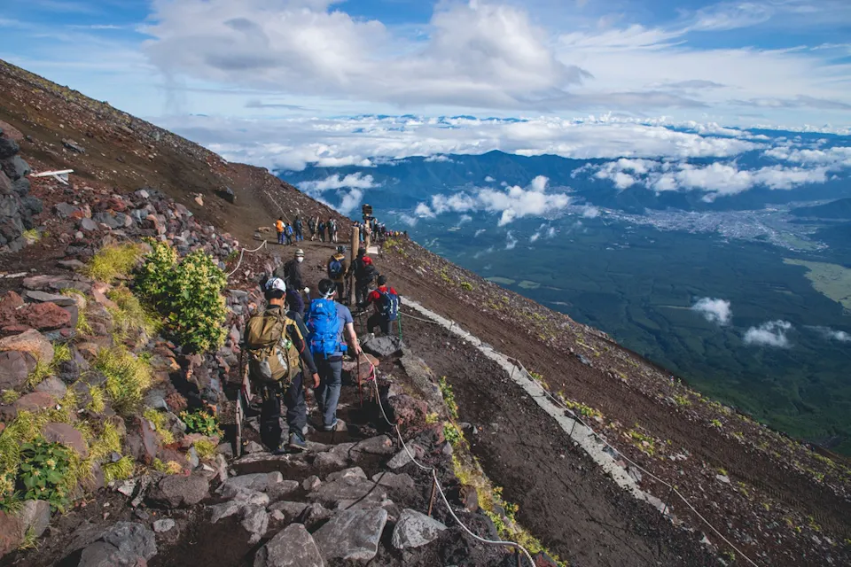 A group of hikers ascending a rocky mountain path. They are dressed in colorful hiking gear with backpacks. The trail is surrounded by rugged terrain and sparse vegetation. In the background, there's a panoramic view of mountains, a valley, and a cloudy sky.