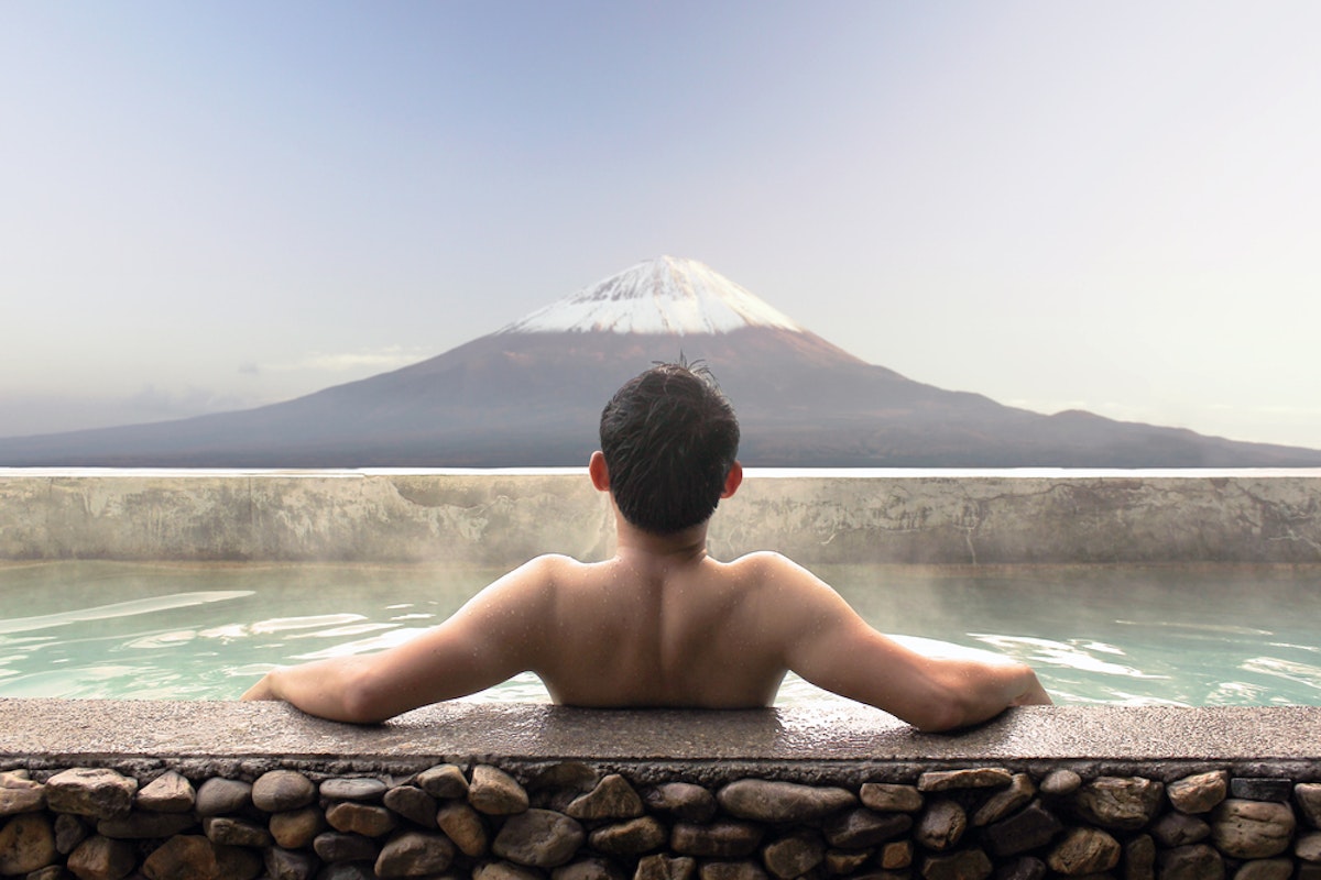 Hot Springs in Mount Fuji A person with dark hair relaxes in an outdoor hot spring, arms outstretched on the stone edge, facing a serene view of a snow-capped mountain under a clear sky. The mountain's reflection is visible in the calm water.