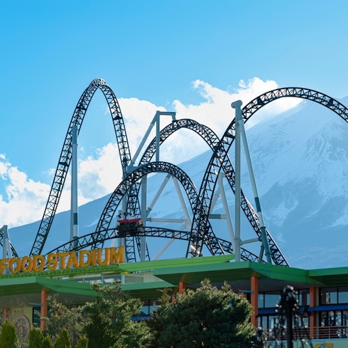 Fuji-Q Highland An amusement park scene with a roller coaster featuring multiple loops and twists in the foreground. "FOOD STADIUM" is written on a building beneath the roller coaster. A snow-capped mountain, possibly Mount Fuji, is visible in the background under a clear blue sky.