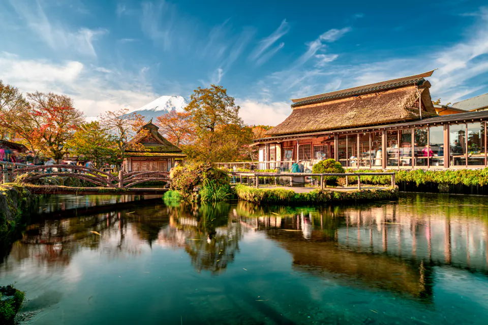 A serene scene features traditional Japanese buildings with thatched roofs by a reflective pond. Autumn foliage surrounds the area, and Mount Fuji is visible in the background under a bright, blue sky with wispy clouds.