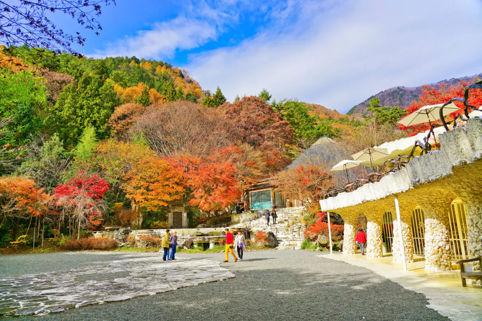 A scenic outdoor area featuring vibrant autumn foliage with reds, oranges, and yellows. People walk along a stone path near a rustic stone building with umbrellas. In the background, forested hills rise under a blue sky with scattered clouds.