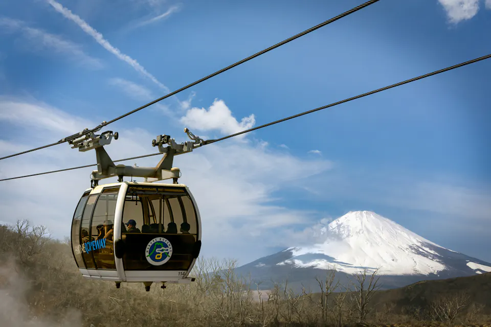 A cable car suspended on a high wire moves across the scene with passengers inside, set against the backdrop of a snow-capped mountain under a clear blue sky. The landscape below shows bare trees and a slightly rugged terrain.
