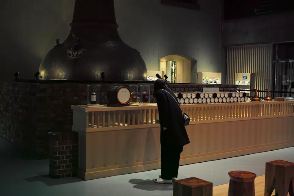 A person in dark clothing leans over a wooden counter examining items in a dimly lit room. In the background, there is a large, vintage distillation apparatus and a brick structure. The setting appears to be an industrial or historical exhibit.