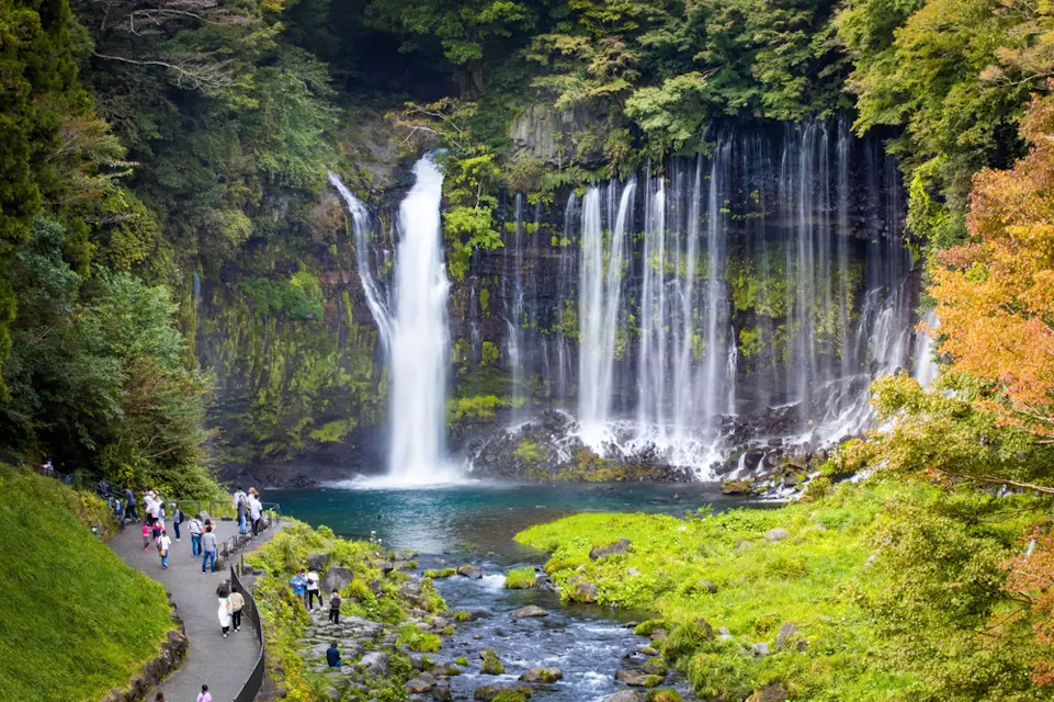 A scenic waterfall cascades over a lush, mossy cliff into a tranquil pool below, surrounded by dense forest. Visitors walk along a paved path on the left, admiring the picturesque view. The area is vibrant with greenery and the autumn colors of the trees.