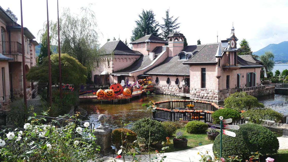 A quaint village with cobblestone buildings surrounded by lush greenery and a serene pond. The area is decorated for Halloween with large pumpkin figures and orange autumn leaves. Mountains and a cloudy sky are visible in the background.