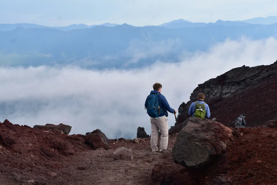 两名背着背包的徒步旅行者沿着一条崎岖的小径步行，靠近一座山的边缘。雾气和云彩笼罩着下面的风景，远处的山脉在地平线上可见。地形崎岖，有大块岩石和红色土壤。
