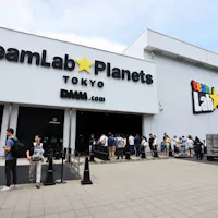 teamLab Planets Tokyo People line up outside the entrance of teamLab Planets Tokyo, a digital art museum. The white building features bold signage for teamLab on the left and DMM.com on the right. The sky is clear, and the ground is paved.