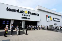 People line up outside the entrance of teamLab Planets Tokyo, a digital art museum. The white building features bold signage for teamLab on the left and DMM.com on the right. The sky is clear, and the ground is paved.
