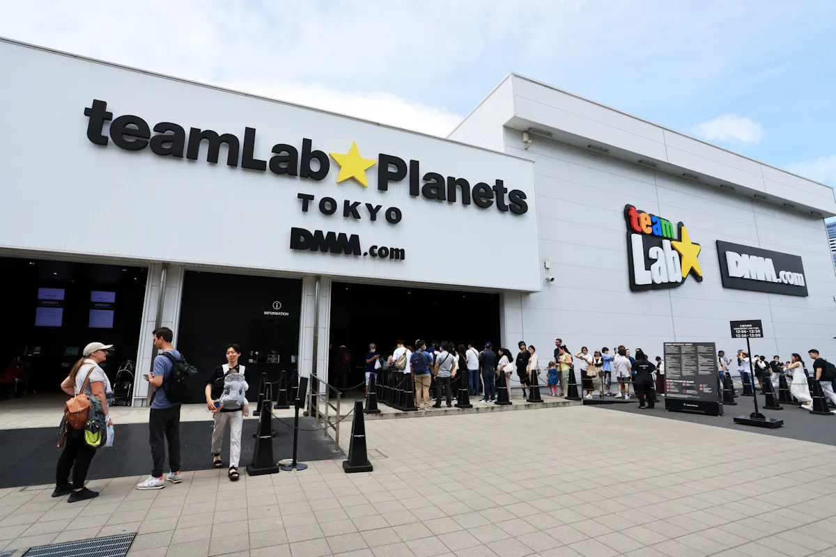 People line up outside the entrance of teamLab Planets Tokyo, a digital art museum. The white building features bold signage for teamLab on the left and DMM.com on the right. The sky is clear, and the ground is paved.