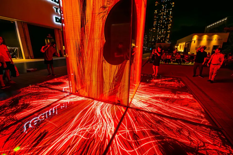 夜间公共空间中的红色和橙色艺术装置被照亮。基座和地面覆盖着复杂的光影图案。人们聚集在周围，欣赏并拍摄这个展览。背景中可见带灯窗户的建筑。