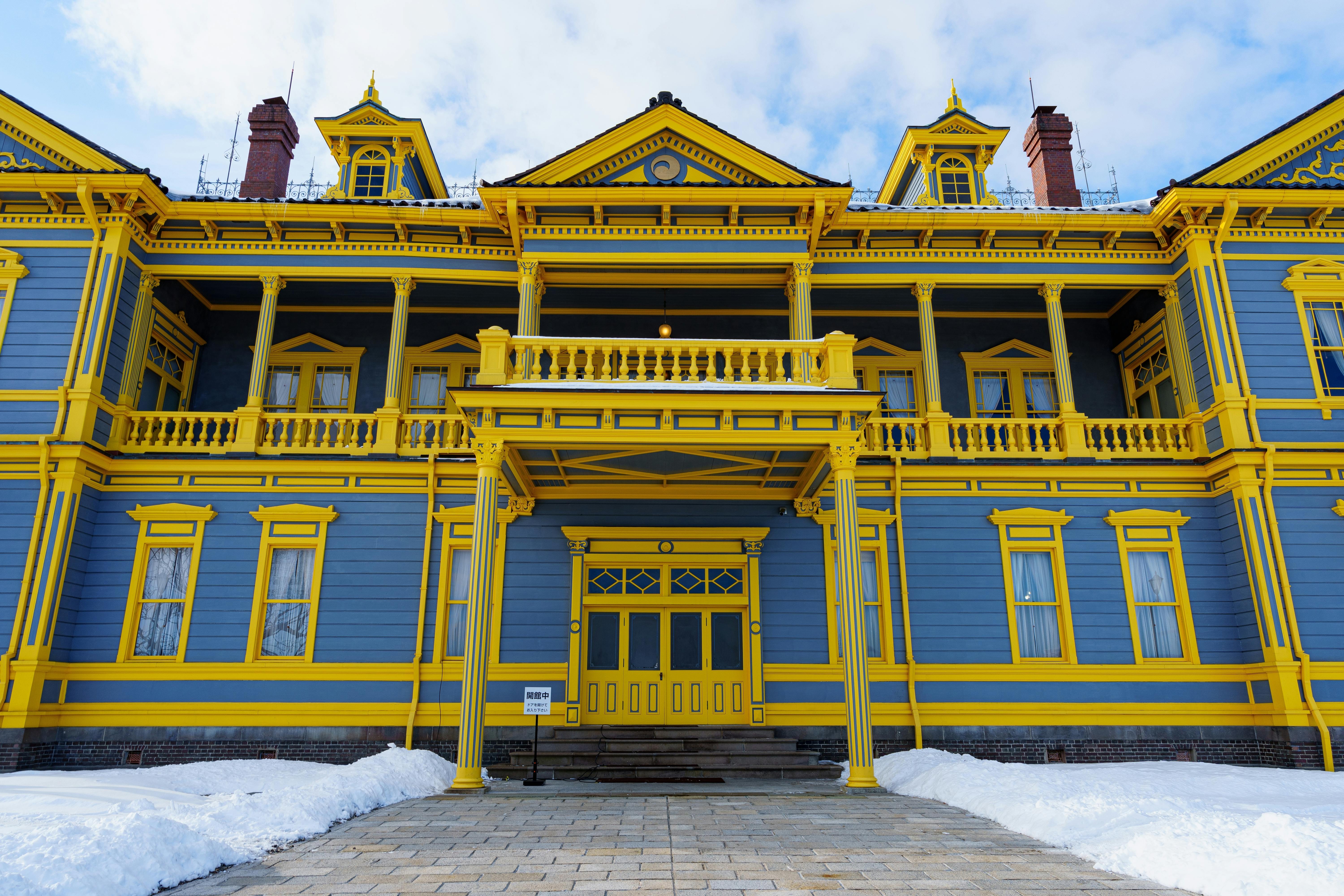 A large, ornate building painted blue with bright yellow trim, featuring columns, balconies, and dormer windows. Snow covers the ground in front, and the sky above is partly cloudy.