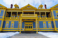A large, ornate building painted blue with bright yellow trim, featuring columns, balconies, and dormer windows. Snow covers the ground in front, and the sky above is partly cloudy.