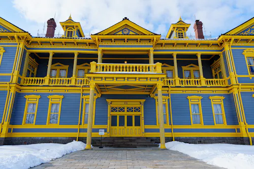 A large, ornate building painted blue with bright yellow trim, featuring columns, balconies, and dormer windows. Snow covers the ground in front, and the sky above is partly cloudy.