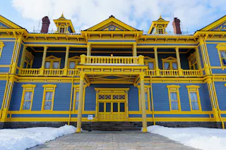 A large, ornate building painted blue with bright yellow trim, featuring columns, balconies, and dormer windows. Snow covers the ground in front, and the sky above is partly cloudy.