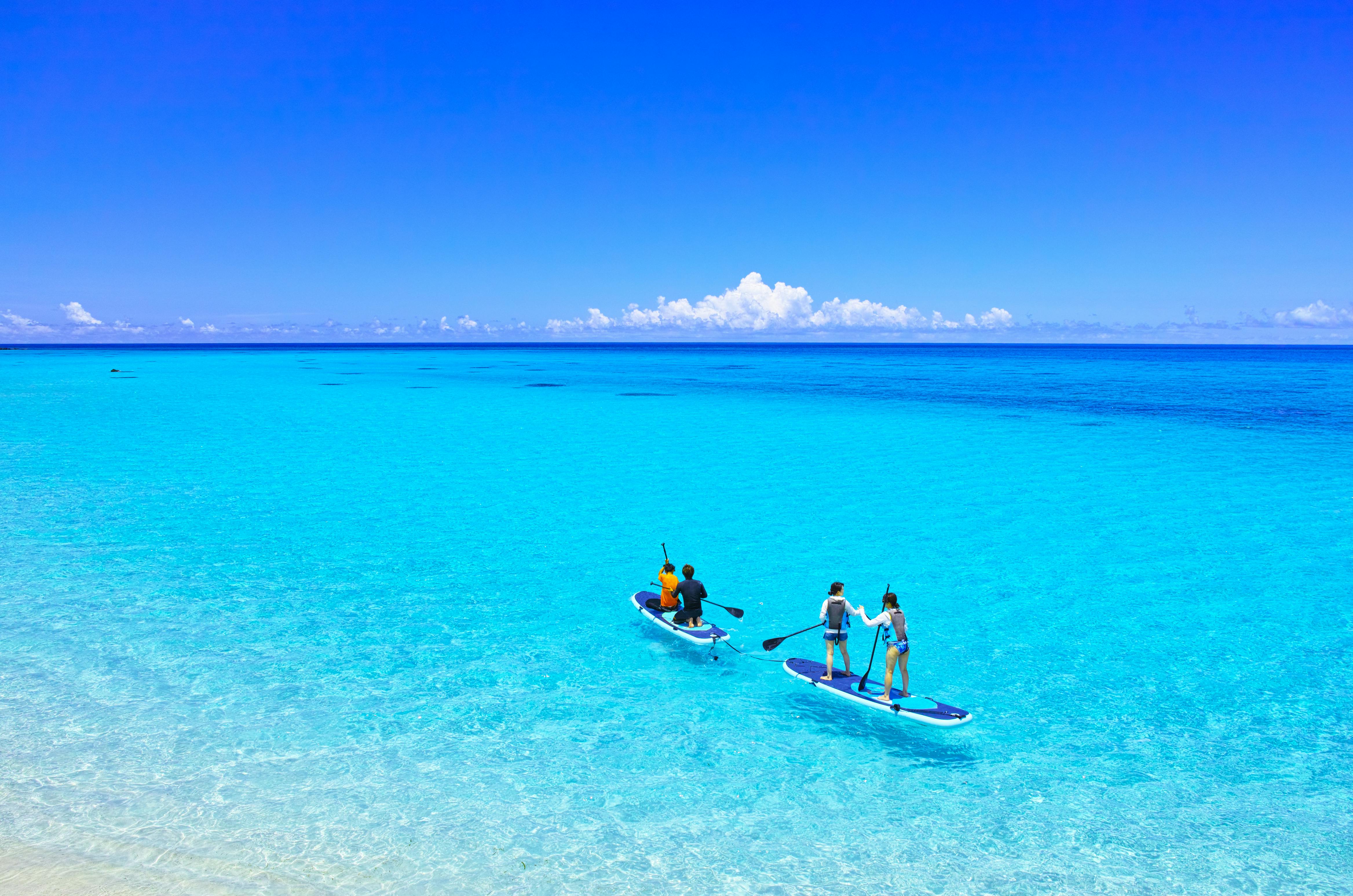 Four people paddleboard on clear, bright turquoise water under a vivid blue sky with scattered clouds. The scene is calm and tropical, with the horizon visible in the distance.