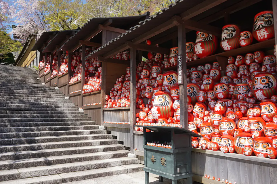 Dharma dedication shelf at Katsuo-ji temple A long outdoor staircase next to a wooden structure filled with numerous red Daruma dolls of varying sizes. The dolls are stacked on shelves and the ground, with some cherry blossom trees visible in the background.
