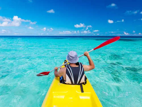 Sea Kayaking A person in a gray hat and life jacket paddles a yellow kayak through clear turquoise water under a bright blue sky with scattered clouds.