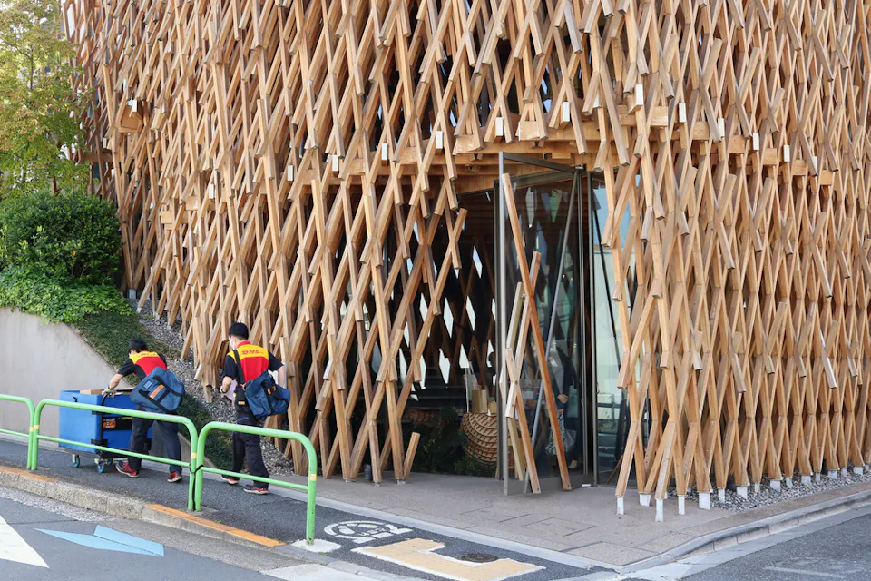 Workers in red and yellow uniforms push blue carts outside a building with a unique facade composed of interlaced wooden beams. The entrance is glass and situated on a corner with green handrails and a ramp leading up to it.