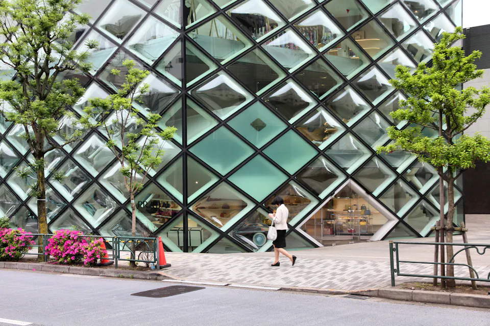A person in business attire walks on a sidewalk in front of a modern glass building with a distinctive diamond-pattern facade. The area features green trees, blooming flowers, and traffic cones near the entrance.