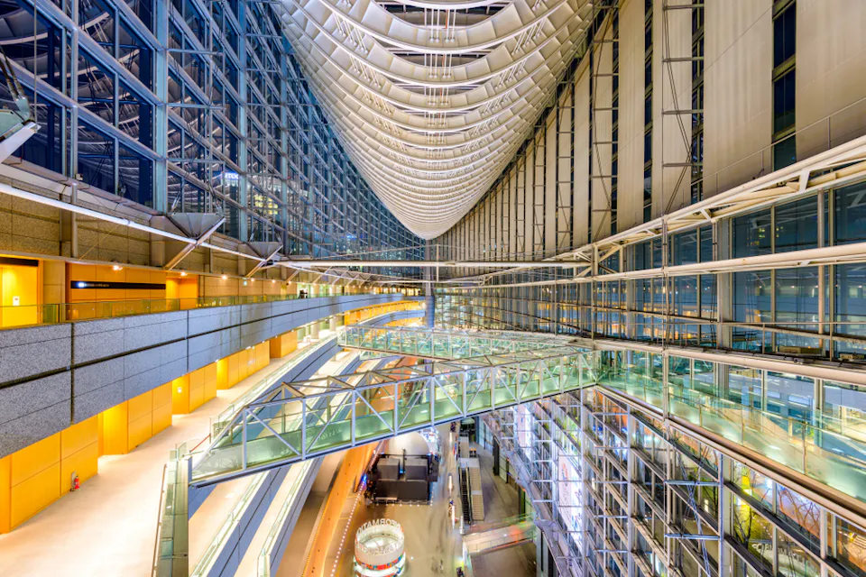A modern architectural interior featuring high glass ceilings and walls with an intricate, wave-like ceiling design. The space has multiple levels, illuminated walkways, and glass bridges connecting areas. The vibrant lighting adds to the contemporary ambiance.