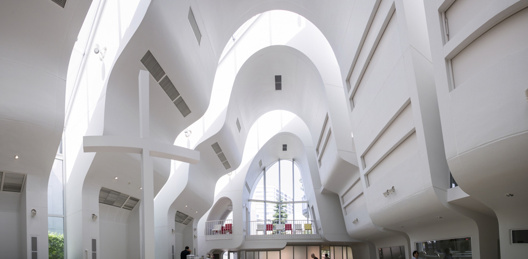 這幅圖片展示了現代建築室內設計，具有流暢、曲線的白色牆壁和高高的天花板。大型窗戶讓自然光進入，創造出通風和寬敞的環境。圖片中出現了人們，賦予設計一種規模感。