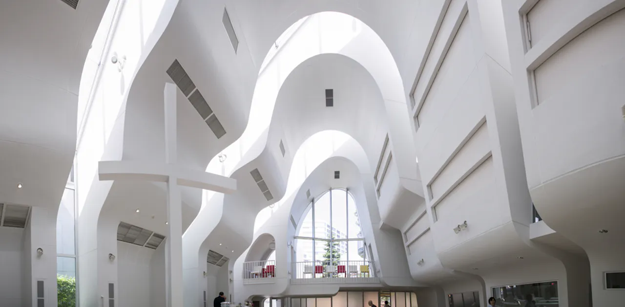 The image shows a modern architectural interior with flowing, curved white walls and high ceilings. Large windows allow natural light to enter, creating an airy and spacious environment. People are present, adding a sense of scale to the design.