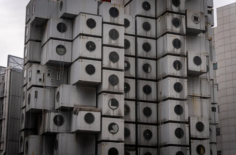 A building with a modular design composed of numerous square units stacked irregularly. Each unit features a large circular window, and some display varying amounts of wear and rust. The sky is overcast, casting a muted light over the structure.