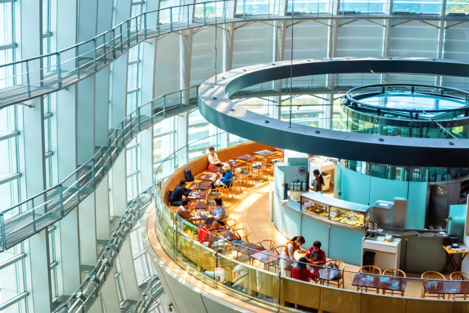A modern, multi-level cafe with glass walls and an open design. People are seated at tables on the circular upper floor, enjoying their meals. The cafe overlooks a sunlit cityscape, with walkways and stairs leading up to the dining area.