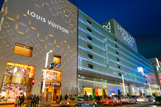A nighttime street view showcasing the bright, illuminated facades of Louis Vuitton and Matsuya Ginza luxury stores in Tokyo, Japan. Crowds of people walk along the sidewalk, and several taxis and vehicles are visible on the street in front.