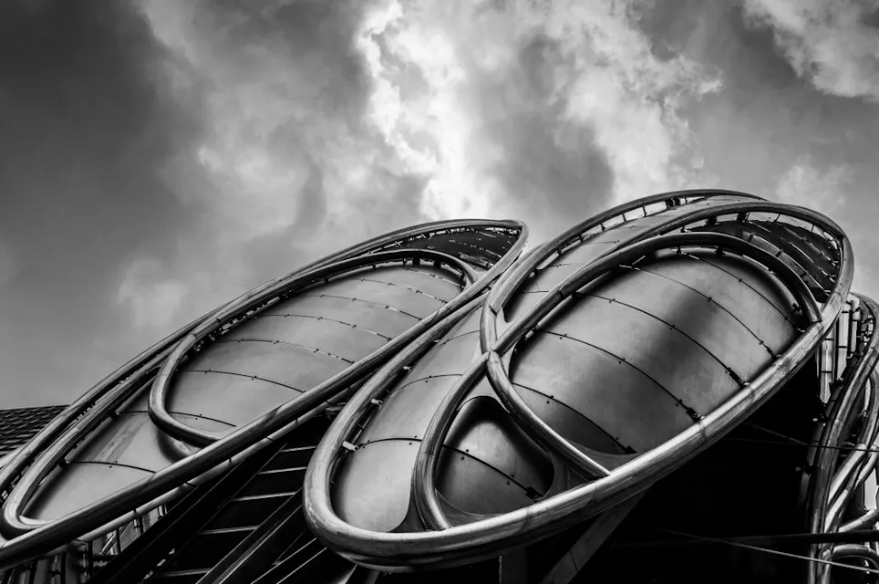 Black and white photo of a modern architectural structure featuring two large, elliptical, metal surfaces with a ribbed design, set against a dramatic sky with clouds. The design is futuristic and sleek.