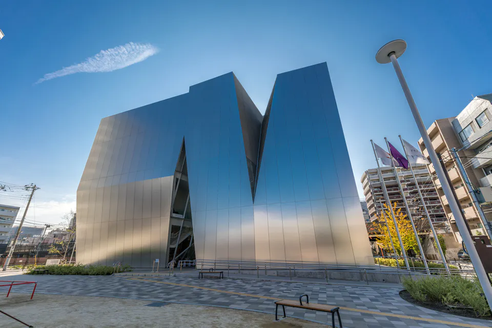 A modern metallic building with geometric shapes stands against a clear blue sky. The building has a reflective surface and an angular design. In the foreground, there are benches, a lamppost, and landscaped areas. Flags and surrounding buildings are visible.