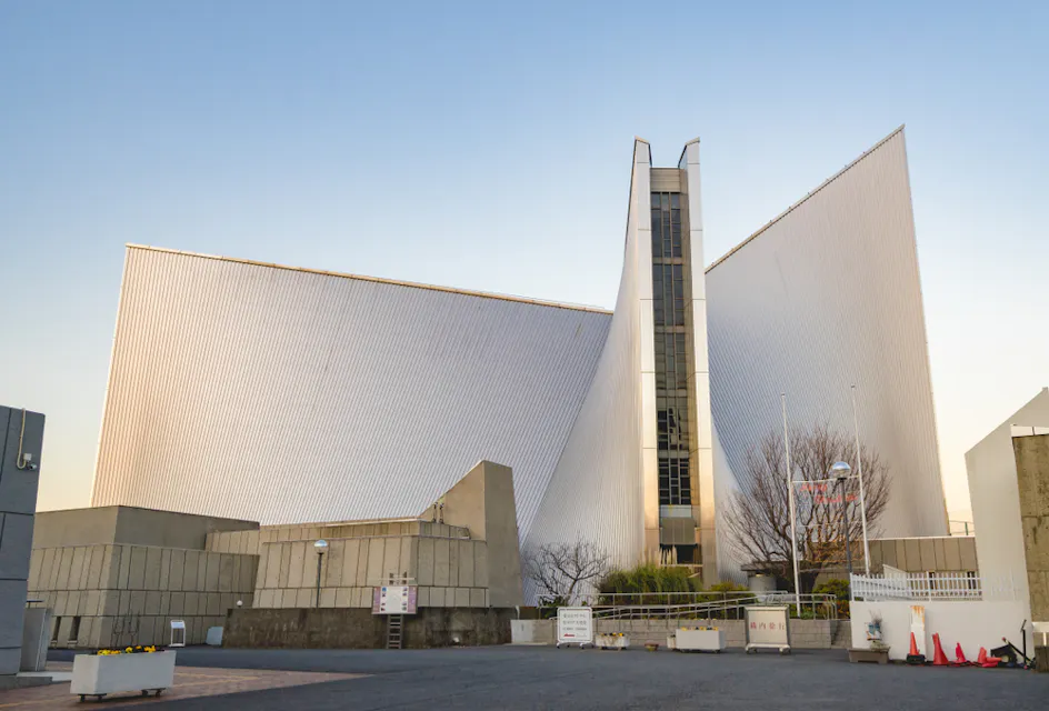 A modern triangular building with sharp angles and a minimalist design. The structure features large, white panels and a central vertical glass element. The foreground has concrete paths, barriers, and a few scattered potted plants. The sky is clear and blue.